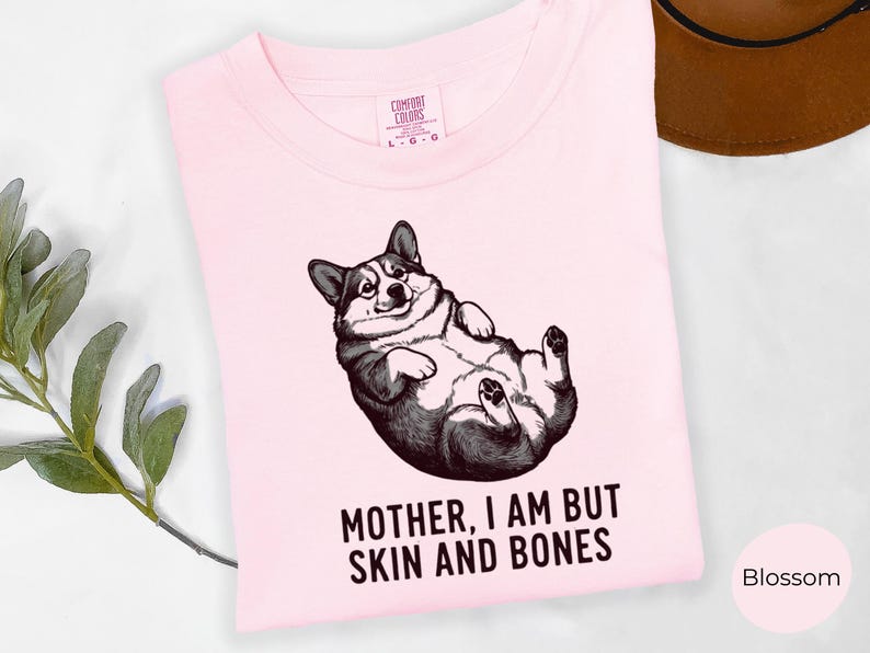 May include: A light pink t-shirt featuring a black and white illustration of a corgi lying on its back. The text "MOTHER, I AM BUT SKIN AND BONES" is printed below the corgi. The shirt is labeled "Blossom".