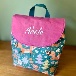 Può includere: Uno zaino in tessuto color verde acqua con un motivo ripetuto di volpi, alberi e fiori. La borsa presenta una patta rosa a tinta unita con il nome Adele ricamato in corsivo bianco. Una maniglia color verde acqua è fissata sulla parte superiore.