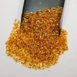 May include: A large pile of small, round, translucent amber-colored gemstones. The gemstones are scattered on a white surface and spilling out of a black tray.