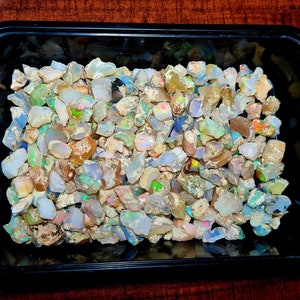 Peut inclure: Un plateau en plastique noir rempli de petites pierres d'opale irisées. Les pierres sont de différentes nuances de blanc, gris et brun avec des reflets de vert, bleu et rouge.