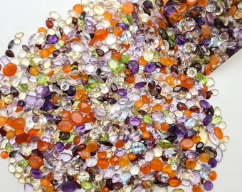 May include: A large assortment of colorful gemstones, including amethyst, citrine, peridot, and carnelian, are scattered on a white surface. The gemstones are various shapes and sizes, and some are in a metal container.
