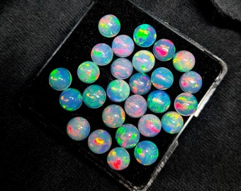 May include: A collection of round, iridescent opal gemstones in various shades of blue, green, and pink. The gemstones are arranged in a black velvet-lined box.