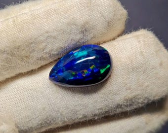 May include: A pear-shaped black opal gemstone with vibrant blue, green, and yellow flashes. The polished stone is held in a white glove, showcasing its iridescent play-of-color. Other opals are visible in the background.