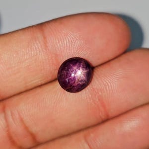 May include: A polished, oval-shaped, dark red gemstone with a six-ray star effect. The gem has a smooth surface. The star effect is visible in the center of the gem. The gem is set against a neutral background.
