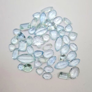 May include: A collection of light blue aquamarine gemstones in various shapes and sizes, including oval, pear, and rectangular cuts.