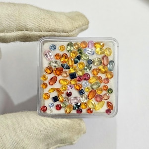 May include: A clear plastic container filled with a variety of colorful gemstones. The gemstones are in various shapes and sizes, including round, oval, and square. Colors include blue, yellow, orange, pink, and green.
