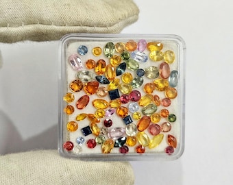 May include: A clear plastic container filled with a variety of colorful gemstones. The gemstones are in various shapes and sizes, including round, oval, and square. Colors include blue, yellow, orange, pink, and green.
