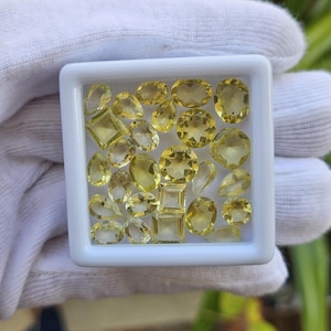 May include: A selection of yellow gemstones in a white tray. The gemstones are cut into different shapes, such as ovals and squares. The gemstones are translucent and are displayed against a white background. The tray is held by a gloved hand.