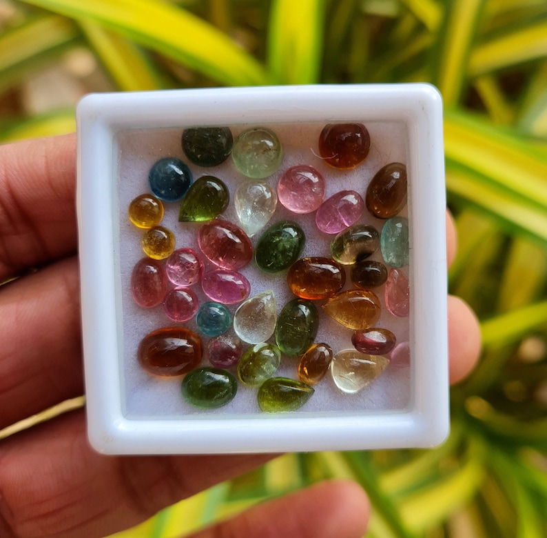May include: A collection of colorful, teardrop-shaped gemstones in a white square container. The gemstones are various shades of green, pink, red, brown, and yellow.