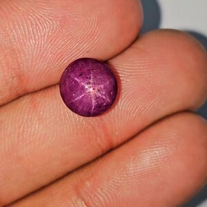 May include: A round, deep magenta gemstone with a six-rayed star pattern. The gem is held in a person's hand, showcasing its polished surface and the star effect created by light reflecting within the stone. The gem is a cabochon cut.