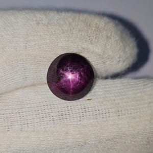 Könnte beinhalten: Ein runder, polierter Edelstein mit tiefvioletter Farbe. Der Edelstein weist einen sechsstrahligen Sterneffekt auf, mit einem helleren, rosavioletten Sternmuster auf seiner Oberfläche. Der Stein ist vor einem weißen Hintergrund platziert.
