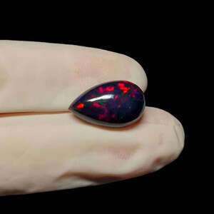 K&ouml;nnte beinhalten: Ein tropfenf&ouml;rmiger schwarzer Opal-Edelstein mit roten und gr&uuml;nen Farbschimmern.