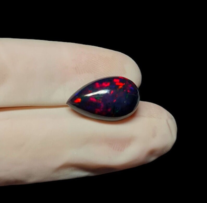 K&ouml;nnte beinhalten: Ein tropfenf&ouml;rmiger schwarzer Opal-Edelstein mit roten und gr&uuml;nen Farbschimmern.