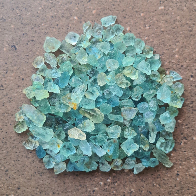May include: A large pile of light blue aquamarine gemstone chips. The chips are irregular in shape and size, with some having a slightly yellow hue.