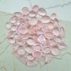 Pode incluir: Uma coleção de gemas de quartzo rosa pálido, cortadas e polidas em forma oval. As pedras estão dispostas em uma pilha sobre uma superfície de mármore branco.