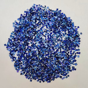 Può includere: Una grande collezione di piccole pietre preziose blu quadrate. Le pietre preziose sono sparse su una superficie bianca.