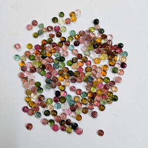 Op de afbeelding: Een verzameling kleine, ronde edelstenen in verschillende kleuren, waaronder roze, groen, geel en bruin. De edelstenen zijn verspreid over een wit oppervlak, waardoor een kleurrijke en gestructureerde weergave ontstaat. Deze edelstenen worden waarschijnlijk gebruikt voor het maken van sieraden.