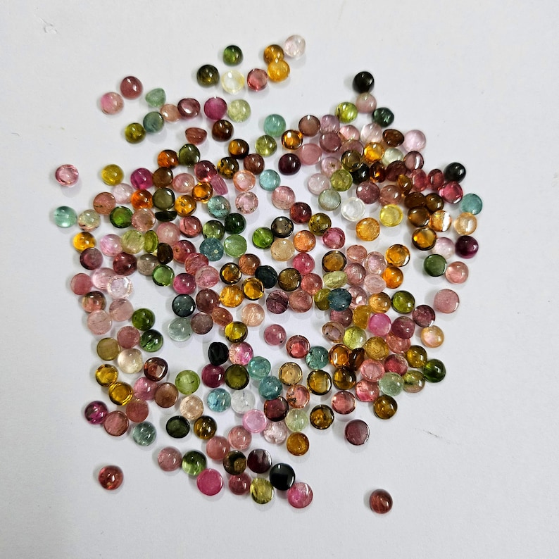 Op de afbeelding: Een verzameling kleine, ronde edelstenen in verschillende kleuren, waaronder roze, groen, geel en bruin. De edelstenen zijn verspreid over een wit oppervlak, waardoor een kleurrijke en gestructureerde weergave ontstaat. Deze edelstenen worden waarschijnlijk gebruikt voor het maken van sieraden.