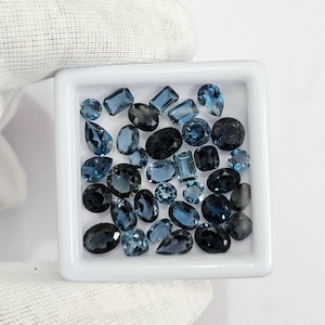 May include: A collection of blue gemstones in various shapes and sizes, including oval, round, and rectangular cuts, are displayed in a white square tray. The gemstones range in color from light to dark blue, with some appearing nearly black. The tray is held by a gloved hand.