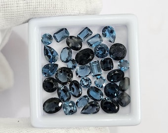May include: A selection of blue gemstones in diverse forms, such as oval, round, and rectangular cuts, are presented in a white square tray. The gemstones vary in colour from light to dark blue, with some appearing almost black. The tray is held by a gloved hand.