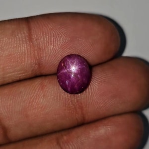 Könnte beinhalten: Ein tiefroter, oval geformter Edelstein mit einem sternartigen Muster auf seiner Oberfläche. Der Edelstein wird in einer Hand gehalten und zeigt seine polierten, reflektierenden Eigenschaften. Die Farbe des Steins ist ein sattes, dunkles Rot mit subtilen Variationen.