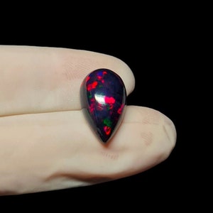 K&ouml;nnte beinhalten: Ein tropfenf&ouml;rmiger schwarzer Opal-Edelstein mit roten und gr&uuml;nen Farbschimmern.