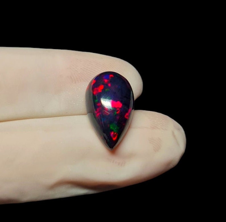 K&ouml;nnte beinhalten: Ein tropfenf&ouml;rmiger schwarzer Opal-Edelstein mit roten und gr&uuml;nen Farbschimmern.
