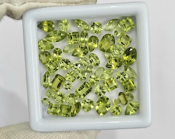 May include: A collection of various-shaped, light green gemstones displayed in a white square tray. The gemstones include oval, pear, square, and round cuts, with a bright, translucent appearance. The tray is held by a gloved hand.
