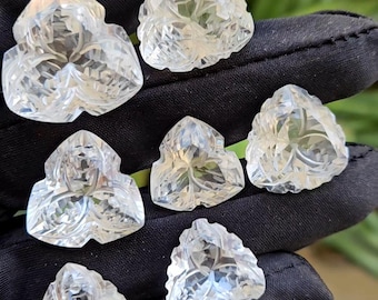 Puede incluir: Una colección de gemas claras y facetadas, cada una con una forma trilobulada única y intrincados diseños florales tallados. Las gemas se sostienen sobre un fondo negro, mostrando su claridad y detalle.