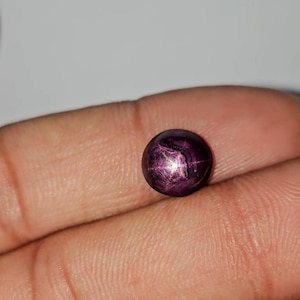 May include: A round, polished gemstone with a deep purple hue. The gem displays a star-like effect across its surface, with lines radiating from a central point. The stone is held in a hand, showcasing its size and color.