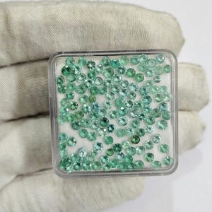May include: A clear plastic container filled with numerous small, round, light green gemstones. The gemstones vary slightly in shade, with some appearing more blue-green. The container is set against a white background.
