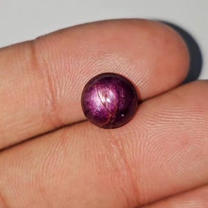 Op de afbeelding: Een ronde, gepolijste edelsteen met een diepe paarsrode kleur. De steen vertoont een ster-effect, met lijnen die vanuit het midden uitstralen. De edelsteen wordt tussen vingers gehouden, wat de grootte en kleur benadrukt. Geschikt voor het maken van sieraden.