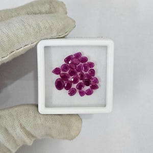 Peut inclure: Un ensemble de pierres précieuses rubis rouge foncé polies de différentes formes, dont des tailles ovales et en poire, présentées dans un récipient carré blanc. Les pierres sont disposées sur une surface blanche, vue de près.