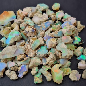 Może przedstawiać: Stosik surowych, naturalnych kamieni szlachetnych z opalu w różnych odcieniach bieli, zieleni, błękitu i pomarańczy. Opale są surowe i mają szorstki, nieregularny kształt.