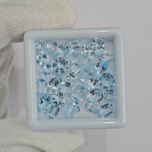 Op de afbeelding: Een verzameling lichtblauwe edelstenen in verschillende vormen, waaronder ovale, ronde en rechthoekige slijpsels, wordt gepresenteerd in een witte vierkante bak. De edelstenen hebben een heldere, sprankelende uitstraling en staan tegen een witte achtergrond.