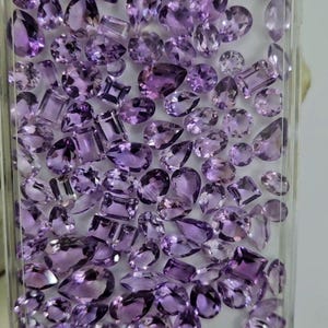 May include: A collection of faceted amethyst gemstones in various shapes, including pear, oval, and square cuts. The gemstones are a deep purple hue and are contained within a clear plastic case, showcasing their brilliance.
