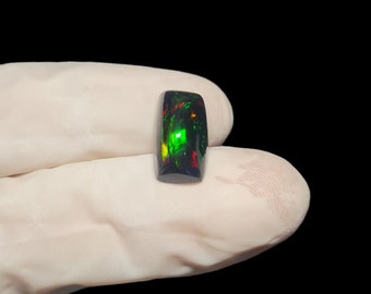 May include: A black opal gemstone with green and red flashes of color. The gemstone is cut into a rectangular shape.