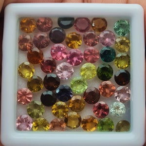 Op de afbeelding: Een verzameling ronde, gefacetteerde edelstenen in verschillende kleuren, waaronder roze, groen, bruin en zwart. De edelstenen zijn gerangschikt in een witte doos.