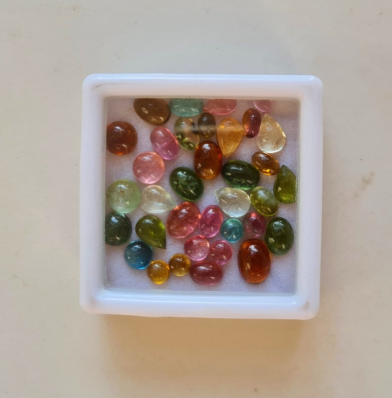 May include: A collection of colorful gemstones in various shapes and sizes, including green, blue, pink, red, and yellow, displayed in a white square container.