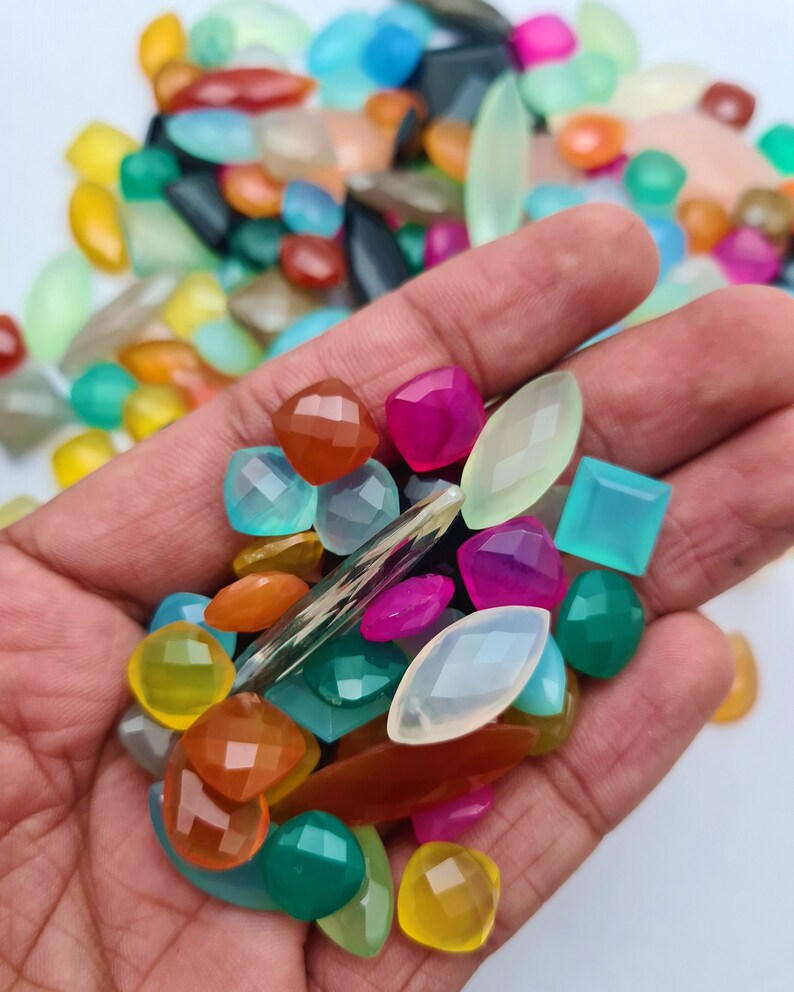 Puede incluir: Una variedad de cabujones de piedras preciosas de colores en diferentes formas y tama&ntilde;os, incluyendo ovalados, redondos, cuadrados y rectangulares. Las piedras preciosas son una mezcla de verde, azul, rosa, naranja, amarillo y blanco.
