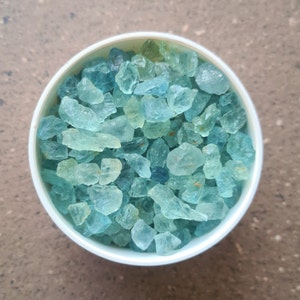May include: A white container filled with aquamarine blue gemstone chips. The chips are of various sizes and shapes.