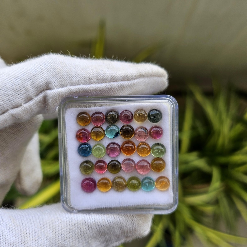 Op de afbeelding: Een verzameling kleurrijke, ronde edelstenen in een doorzichtige plastic doos. De edelstenen zijn in verschillende kleuren, waaronder rood, roze, groen, blauw en geel. De doos wordt vastgehouden door een gehandschoende hand, met een vage groene achtergrond.
