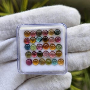 Op de afbeelding: Een doorzichtige plastic doos gevuld met een verscheidenheid aan kleine, ronde edelstenen. De edelstenen zijn een mix van kleuren, waaronder roze, groen, geel, blauw en bruin. De doos ligt op een witte ondergrond.
