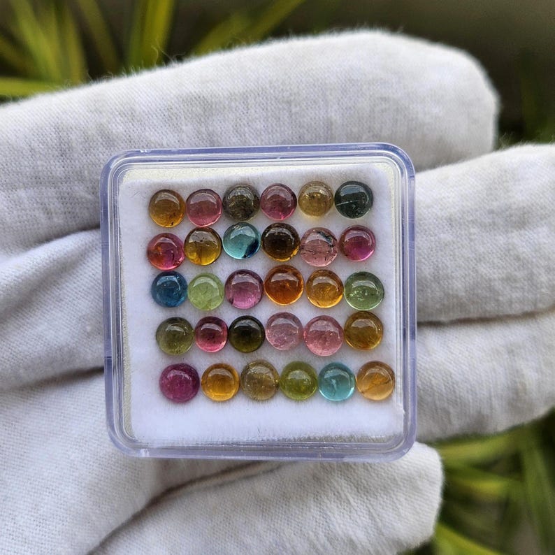 Op de afbeelding: Een doorzichtige plastic doos gevuld met een verscheidenheid aan kleine, ronde edelstenen. De edelstenen zijn een mix van kleuren, waaronder roze, groen, geel, blauw en bruin. De doos ligt op een witte ondergrond.