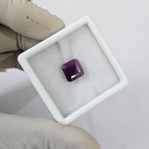 Può includere: Una gemma sfaccettata, viola scuro, taglio smeraldo, presentata in un portagioie quadrato bianco. La gemma ha una forma rettangolare con angoli tagliati, su sfondo bianco.