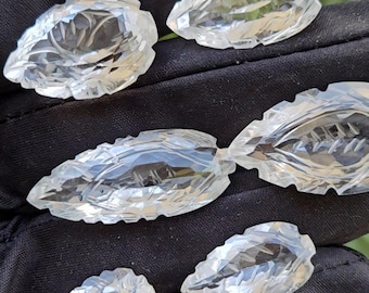Puede incluir: Una colección de tallas de cristal transparentes en forma de hojas. Los cristales están facetados, captando la luz y mostrando detalles intrincados. Las hojas varían en tamaño y se muestran sobre un fondo oscuro.