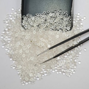 以下が含まれることがあります： 白い表面に散らばった、小さな透明な丸いガラスビーズの山。その上に黒いピンセットが置かれている。