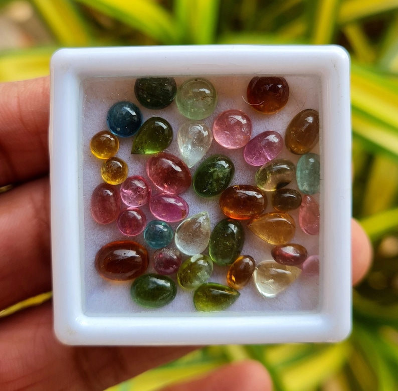 May include: A white box filled with a variety of small, colorful gemstones. The gemstones are in various shades of green, pink, red, brown, and yellow.