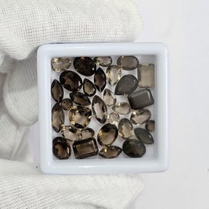 Può includere: Una collezione di gemme sfaccettate di varie forme e dimensioni, tra cui ovali, a goccia e rettangolari. Le gemme sono di diverse tonalità di marrone e fumé, esposte in un vassoio quadrato bianco.