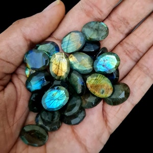 Pode incluir: Uma coleção de gemas de labradorita de forma oval em tons de verde, azul e dourado. As pedras têm um brilho iridescente e são exibidas em uma mão.